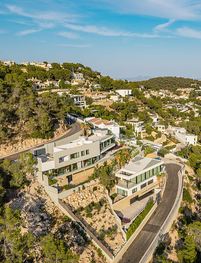 Aerial drone shot of a white contemporary hillside villa surrounded by pine trees, with a winding road and coastal horizon.