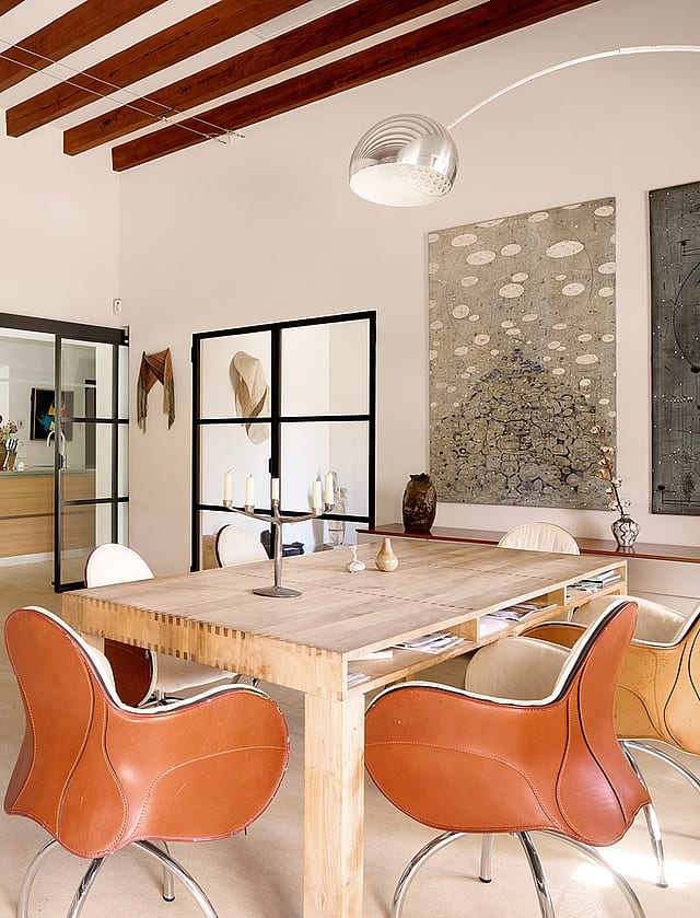 Dining area featuring a rustic wooden table, leather bucket chairs, and exposed timber ceiling beams in a spacious white room.