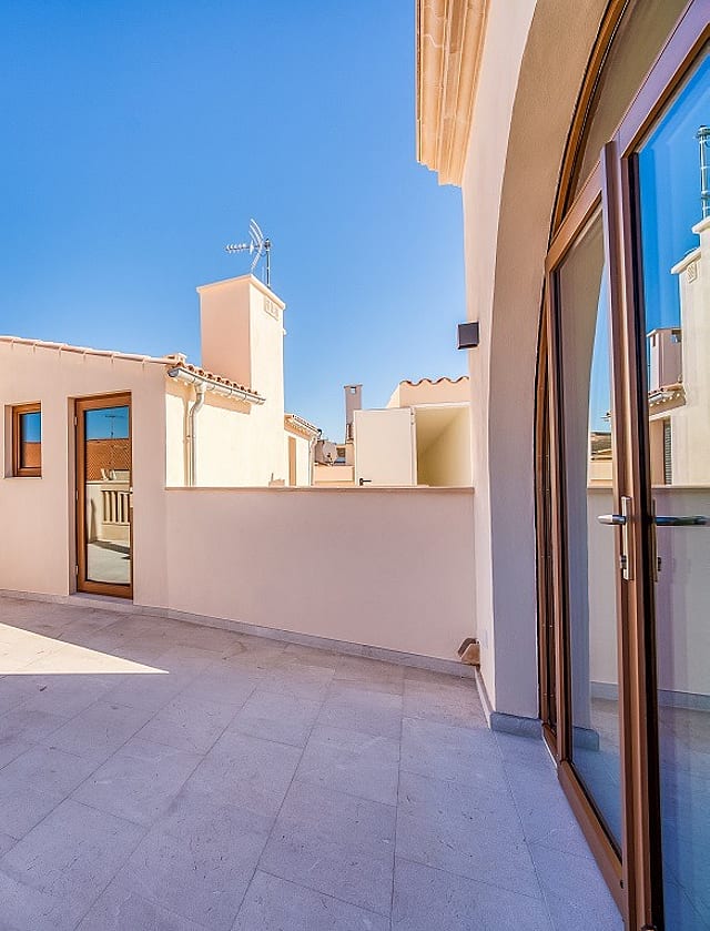An open stone-paved rooftop terrace with light-colored walls and glass doors under a clear blue sky, overlooking city buildings.