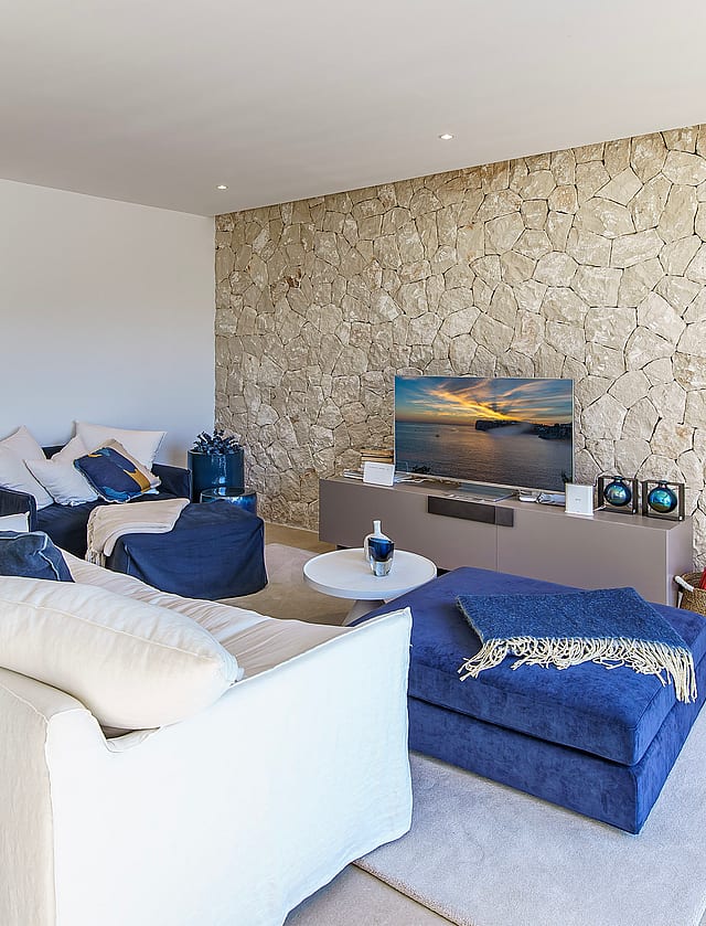 Modern living room featuring a textured natural stone wall, white sofa, blue armchairs, and neutral tiled flooring.