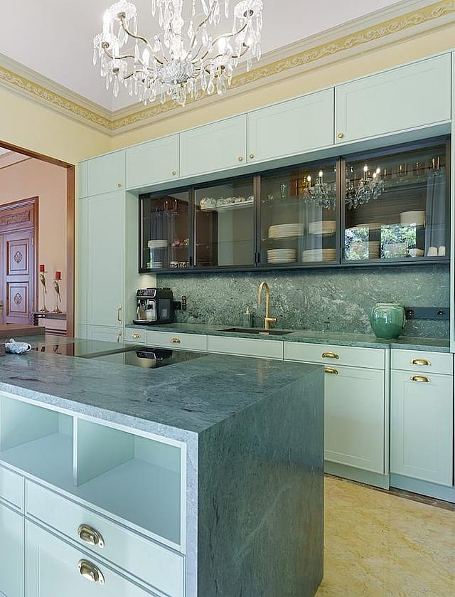 Modern kitchen with sage green cabinets, a dark grey stone island, and bar stools, leading to a dining area with a chandelier.
