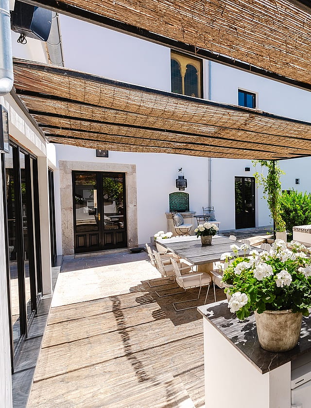 An outdoor kitchen with stone counters and a sink sits under a woven reed canopy, adjoining a paved terrace and garden.