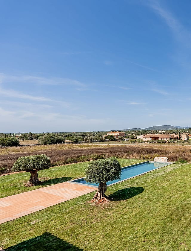 An elevated view over a green lawn and rectangular pool toward distant mountains in the Santanyí countryside.