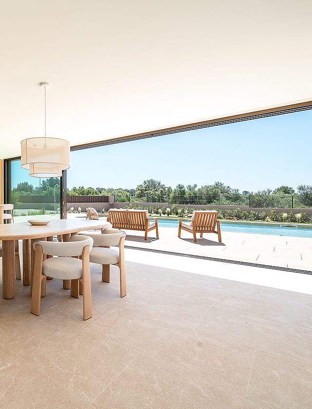 Bright dining area with oval wooden table and cream upholstered chairs, large sliding doors opening to an outdoor pool deck.