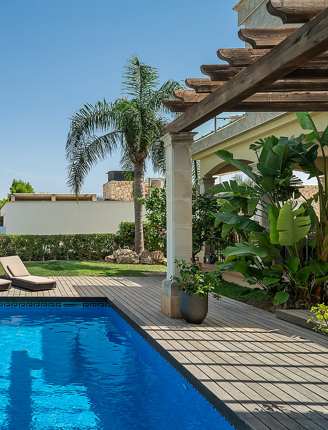 Rectangular swimming pool with blue water next to a timber deck, two sun loungers, and a shaded pergola in a sunny garden.
