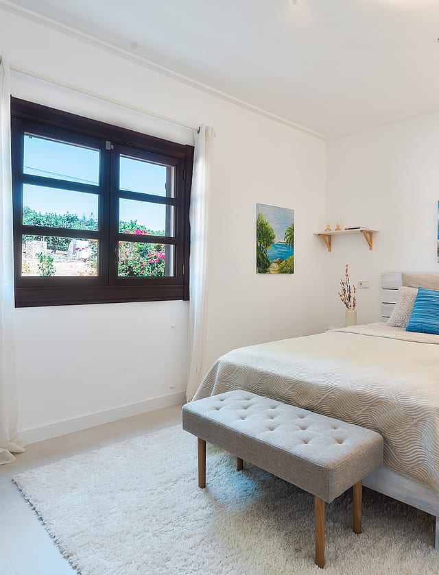 Dormitorio con cama de matrimonio, alfombra de texturas y marco de ventana de madera oscura hacia el verde.
