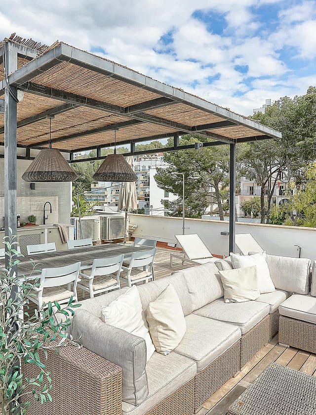 A rooftop terrace featuring a wicker sectional sofa, dining area, and outdoor kitchen under a wooden pergola.