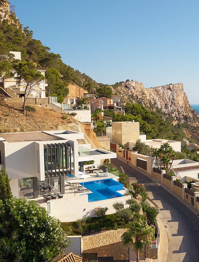 Aerial view of a white cliffside villa with a blue swimming pool, situated along a coastal road overlooking the Mediterranean.