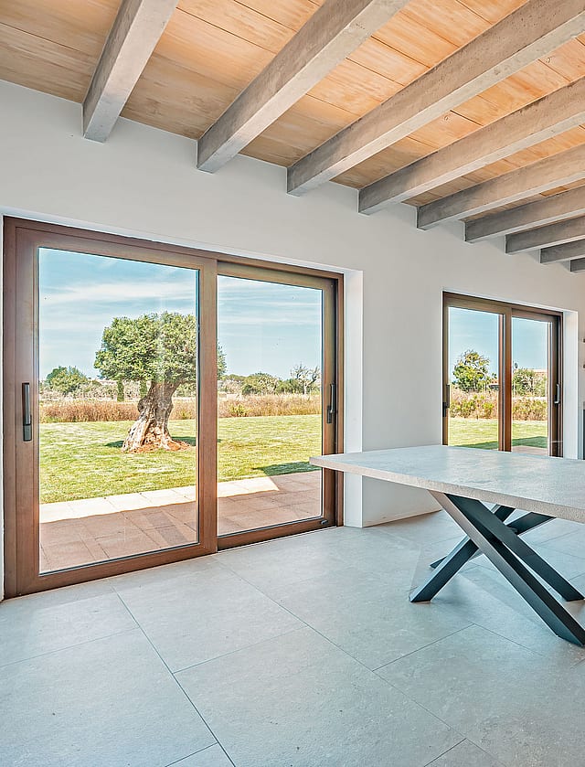 Dining area with a stone table and dark metal legs, exposed wooden ceiling beams, and large glass doors overlooking a rural landscape.