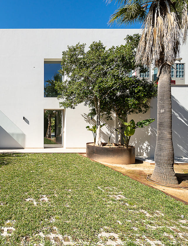 Modern white minimalist villa exterior featuring a flat roof, large rectangular windows, and a manicured green lawn with mature trees.