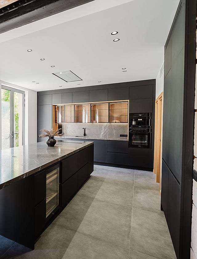 Modern open-plan kitchen in an apartment featuring a large dark stone island with waterfall edges, matte black cabinetry, and exposed wooden ceiling beams.