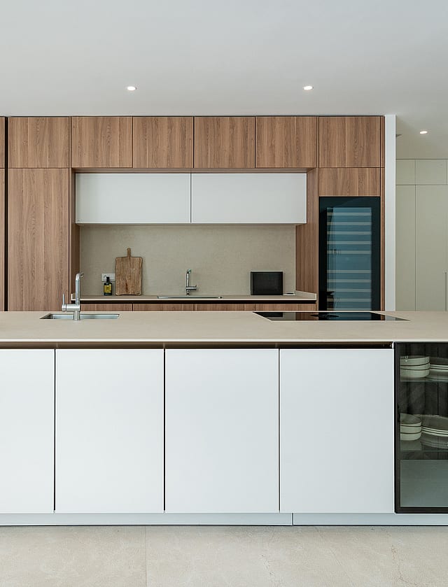 White handleless Siematic kitchen island with stone worktop, integrated sink and glass-fronted crockery display, floor-to-ceiling oak-veneer cabinetry and Gaggenau ovens behind