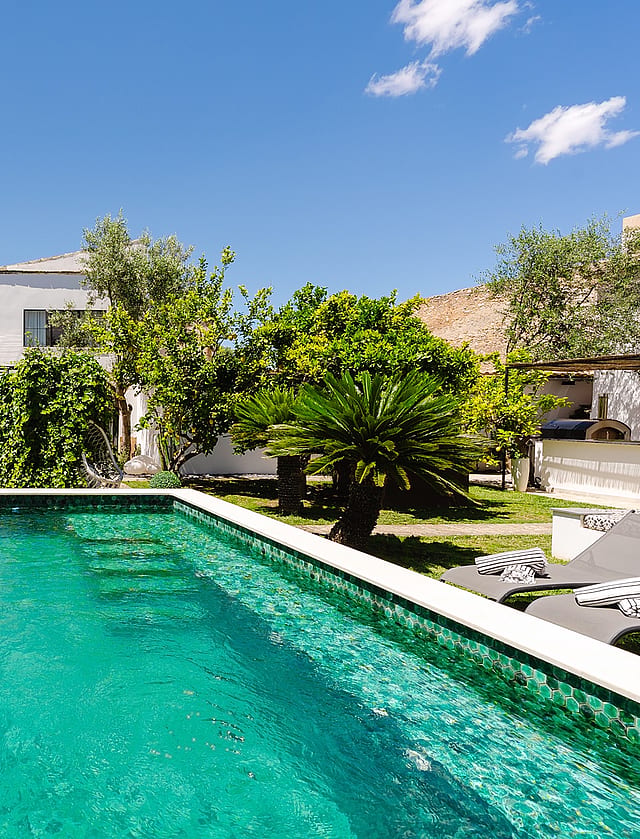 A rectangular swimming pool with turquoise mosaic tiles set within a lush, walled garden in Sencelles, featuring mature trees.
