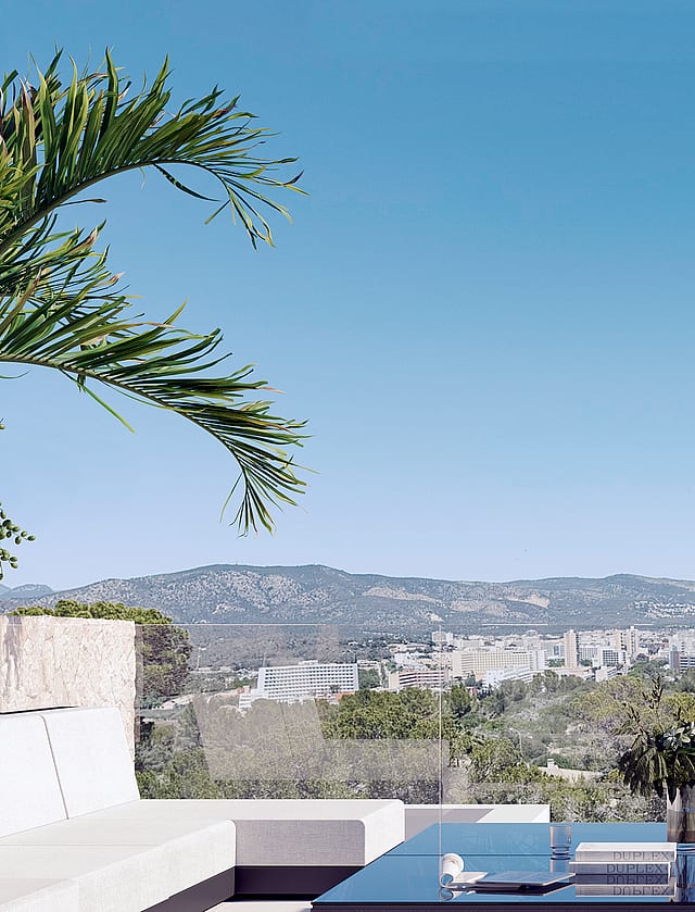 An outdoor terrace with white minimalist sofa and glass-topped table overlooking a valley, cityscape, and mountain range.