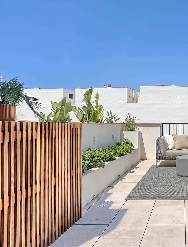 A spacious rooftop terrace featuring a modular outdoor sofa set on a textured rug, bordered by a wooden screen and stone walls.