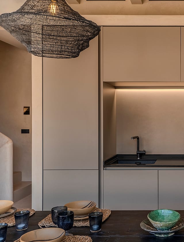 A minimalist kitchen wall featuring matte beige cabinetry, a dark countertop, integrated oven, and a sculptural textured pendant light.