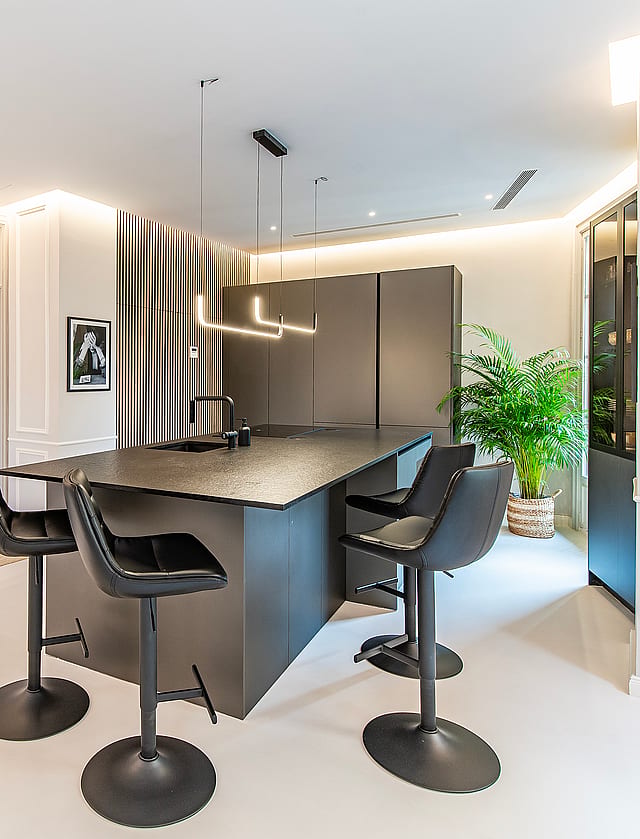 Modern kitchen featuring a dark grey central island, black swivel stools, recessed ceiling lighting, and panelled walls.