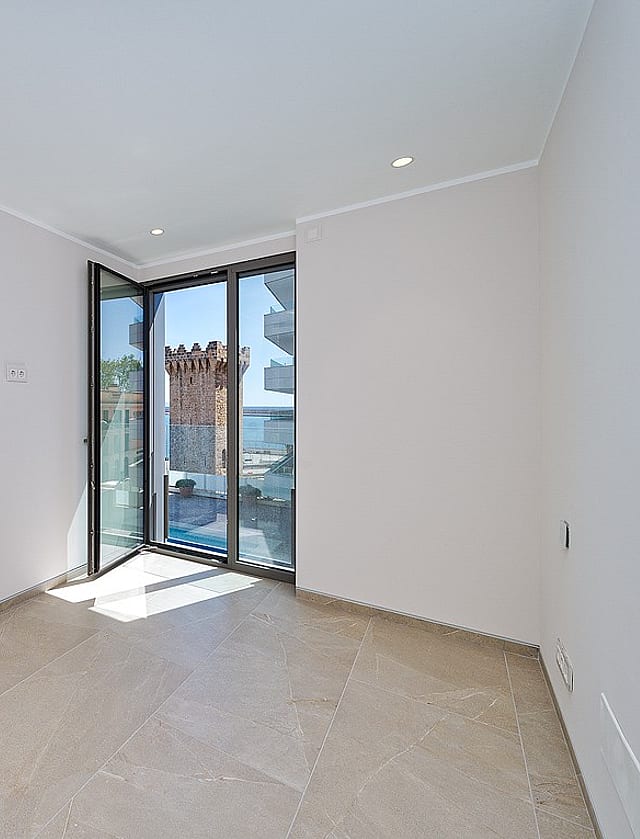 Bright unfurnished bedroom with pivot glass door opening to balcony, Torre de Paraires and harbour visible beyond
