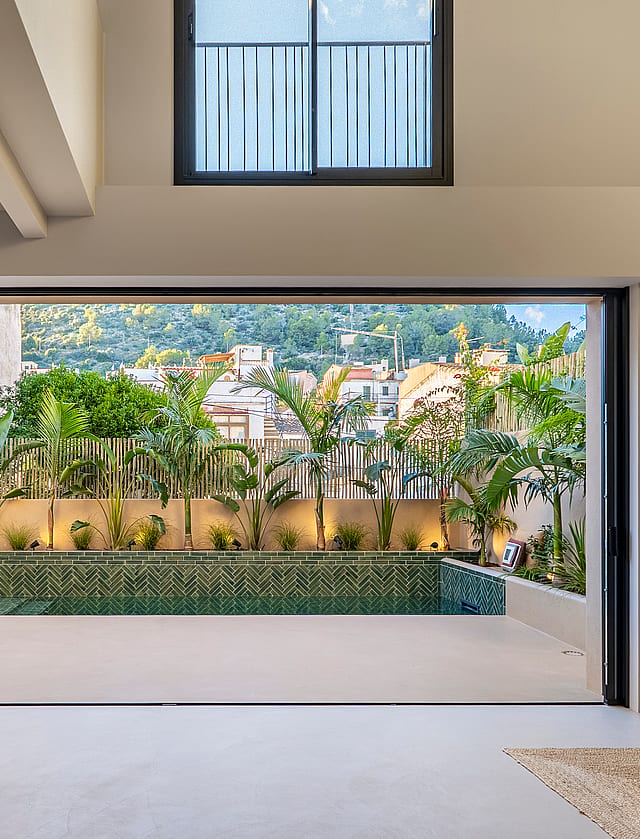 Interior view through open sliding doors to a terrace with a green tiled plunge pool and lush perimeter planting.