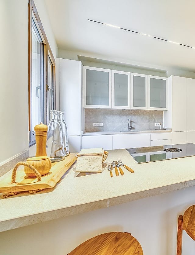 Modern kitchen with white cabinetry, integrated appliances, and a light stone island with wooden bar stools in the foreground.