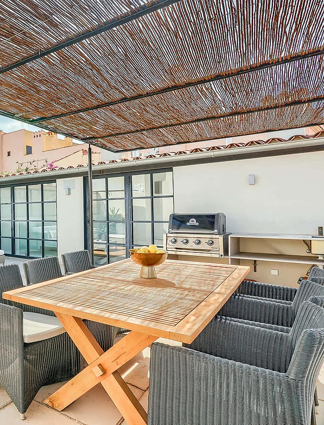 A spacious outdoor terrace with a wooden dining table, wicker chairs, and a shaded lounge area against a city backdrop.
