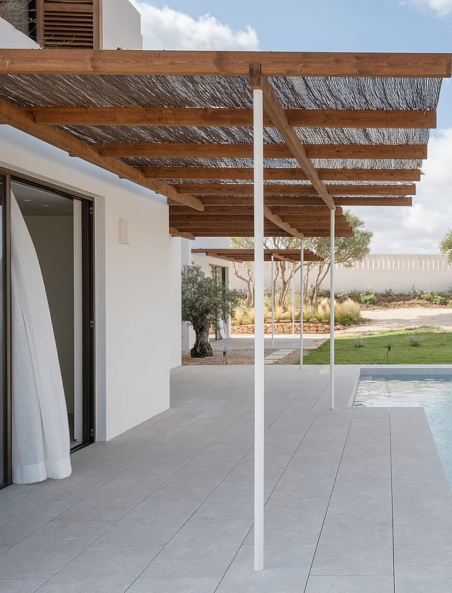Modern pool terrace featuring white stone tiling and a timber pergola with reed shading next to a garden lawn.