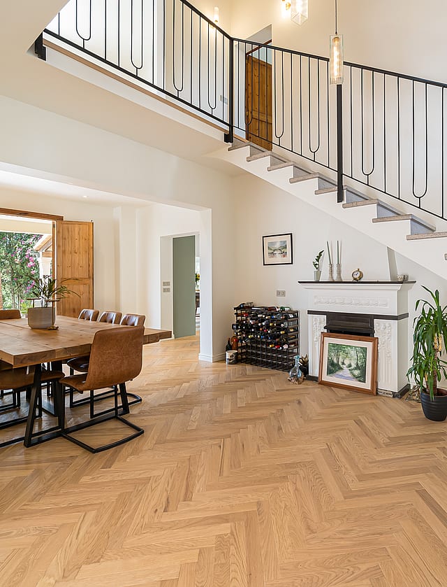 Sunlit living space with oak chevron floors, a black iron staircase, and a dining table near open wooden shutters.