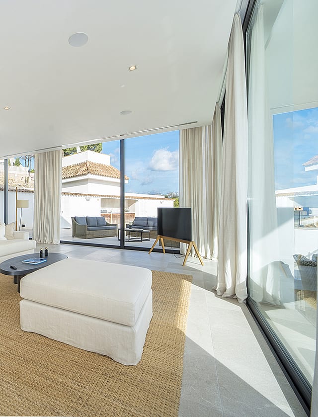 Bright living room with white L-shaped sofa, matching ottoman, woven area rug, and floor-to-ceiling glass sliding doors.