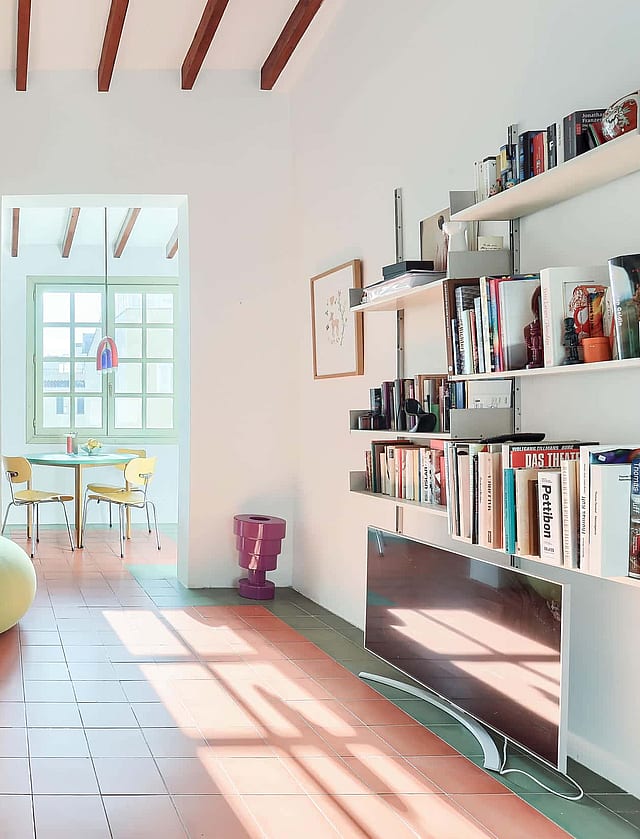 A bright living area featuring terracotta floor tiles, exposed wooden ceiling beams, a sofa with a striped cushion, and a round lime green ottoman, looking through to a dining area.