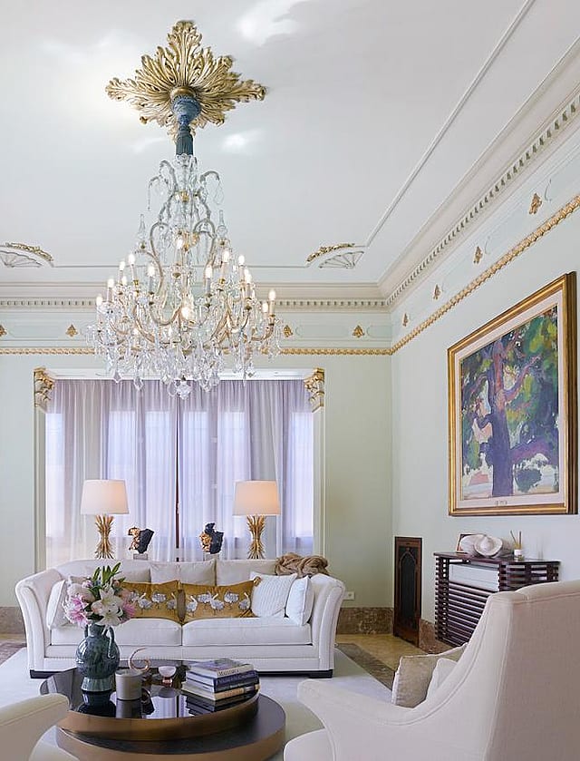 A formal living room with high ceilings, decorative gilded molding, and a crystal chandelier above a white sofa.