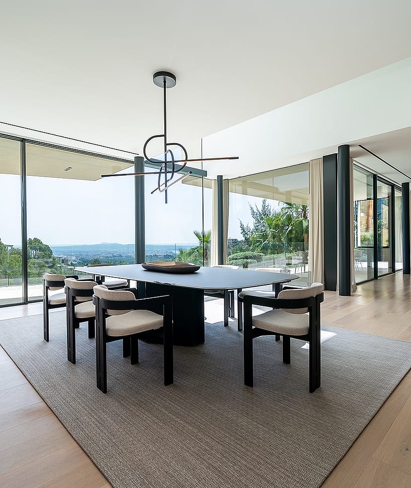 Dining room with an oval table and six chairs on a textured rug, floor-to-ceiling glass walls, and oak wood flooring.