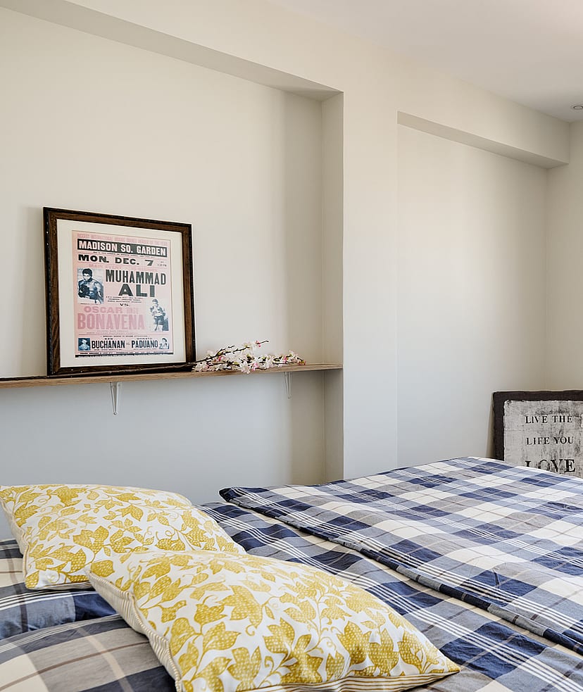 A bedroom featuring a bed with a blue plaid quilt, a floating wooden shelf with framed artwork, and a window overlooking city buildings.