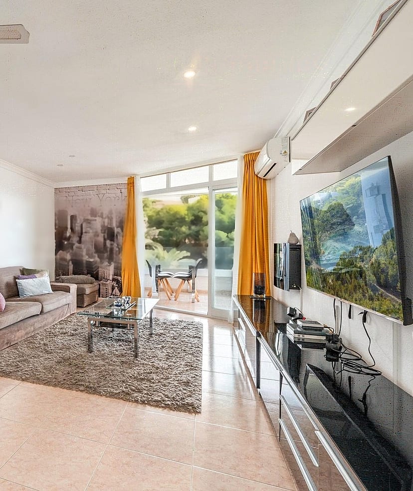Wide-angle view of a living room featuring a plush sofa, high-gloss TV unit, and floor-to-ceiling glass doors opening to a terrace.