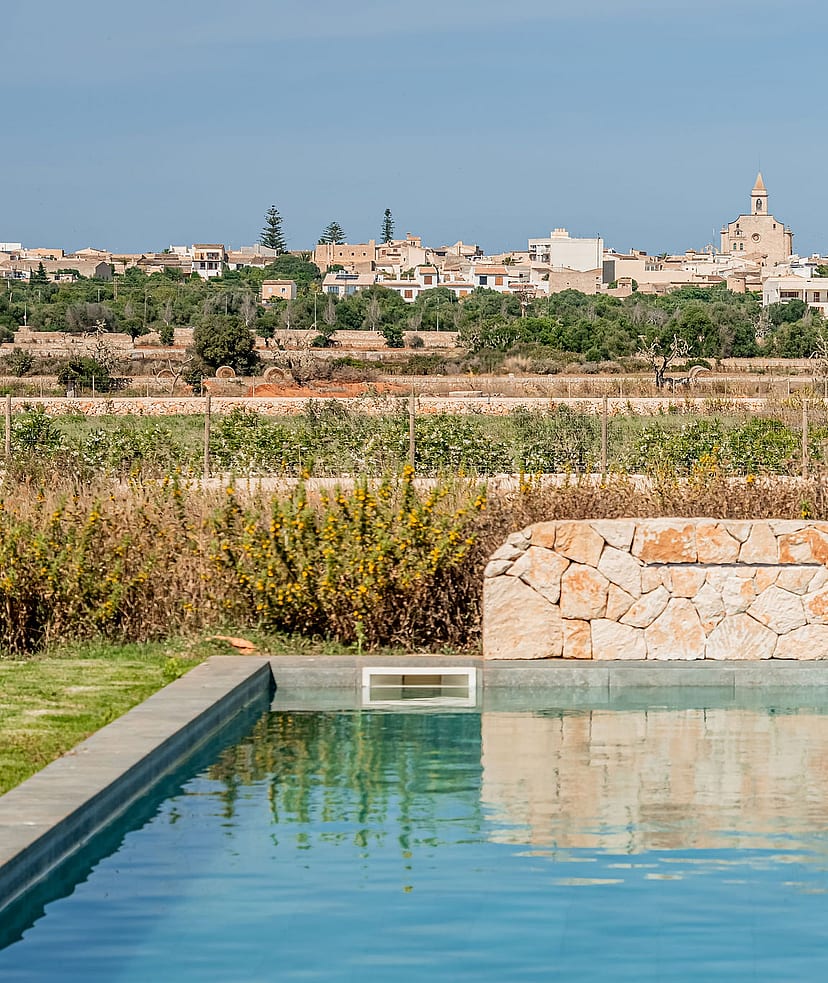 A rectangular swimming pool featuring a natural stone waterfall wall with the village of Santanyí in the background.