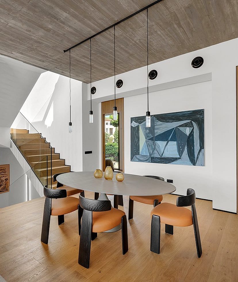 Dining area with oval table and wooden chairs, adjacent to a wooden staircase under a raw concrete ceiling.
