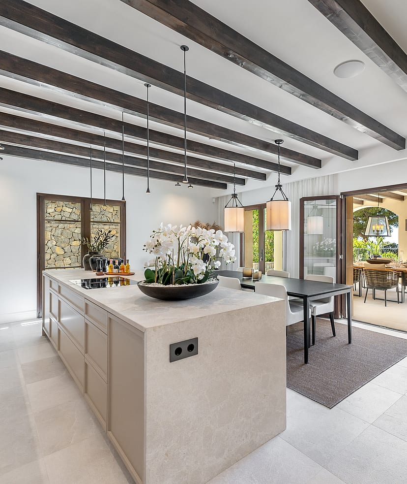 A wide stone kitchen island and dark ceiling beams in an open-plan room opening onto a sunlit outdoor dining terrace.