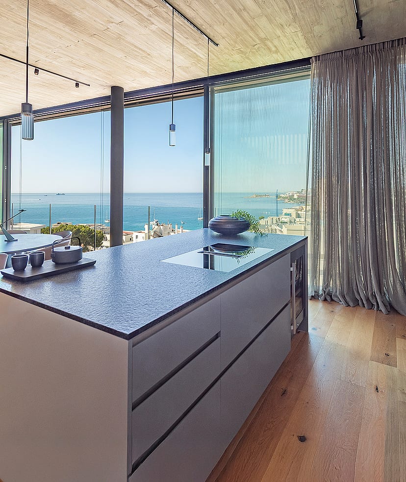Modern kitchen island with dark stone countertop, light wood flooring, and floor-to-ceiling glass windows overlooking the sea.