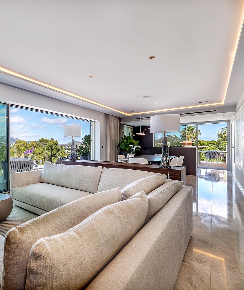 A contemporary open-plan living room featuring a large beige sectional sofa, polished stone floors, and floor-to-ceiling glass sliding doors opening to a lush garden view.