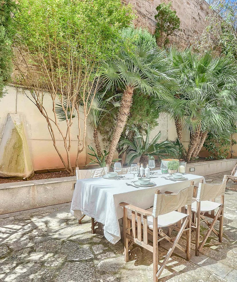 An outdoor dining table with white tablecloth and director chairs set on a gravel terrace shaded by mature trees.