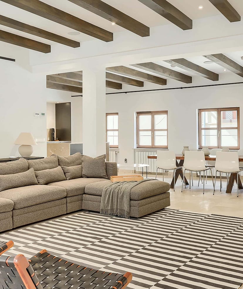 A spacious living room featuring a large patterned sofa, exposed dark wood ceiling beams, and a dining area in the background.