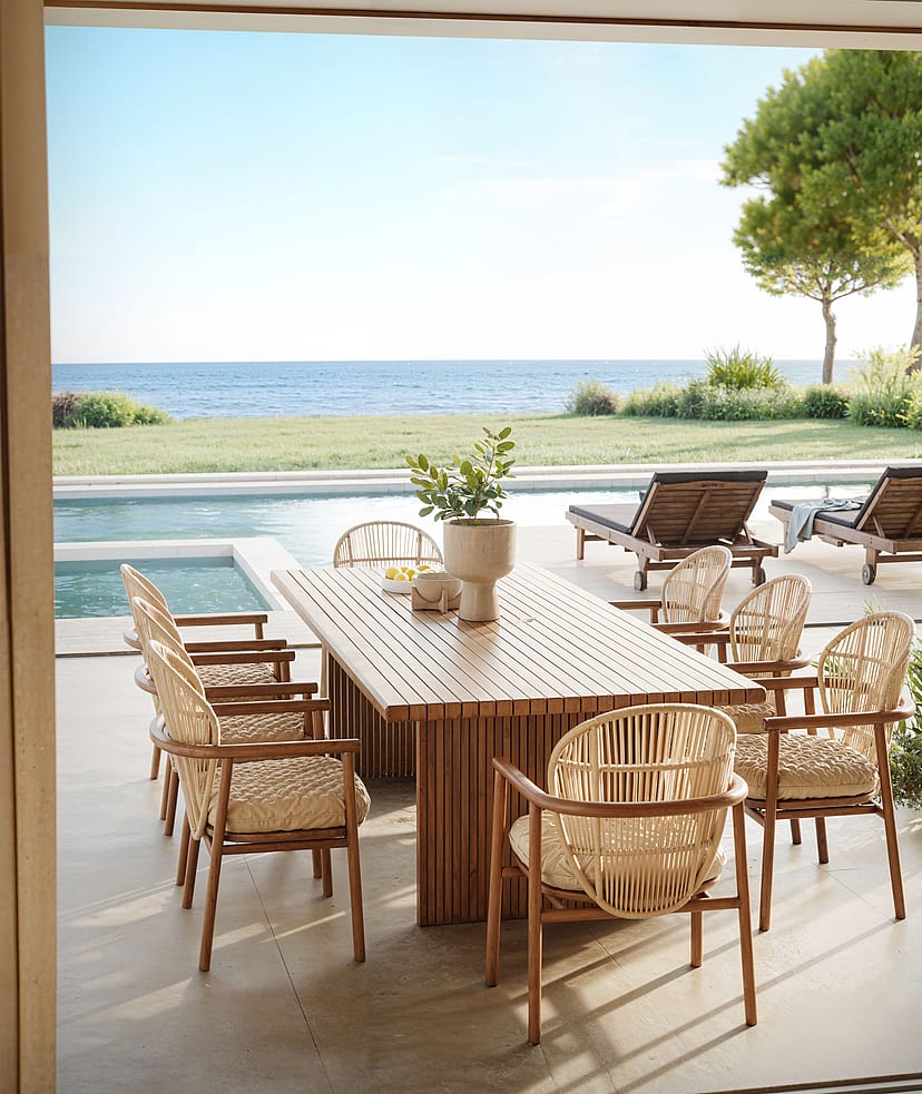 A slatted wood outdoor dining table with wicker chairs on a covered terrace, facing a swimming pool and the Mediterranean sea.