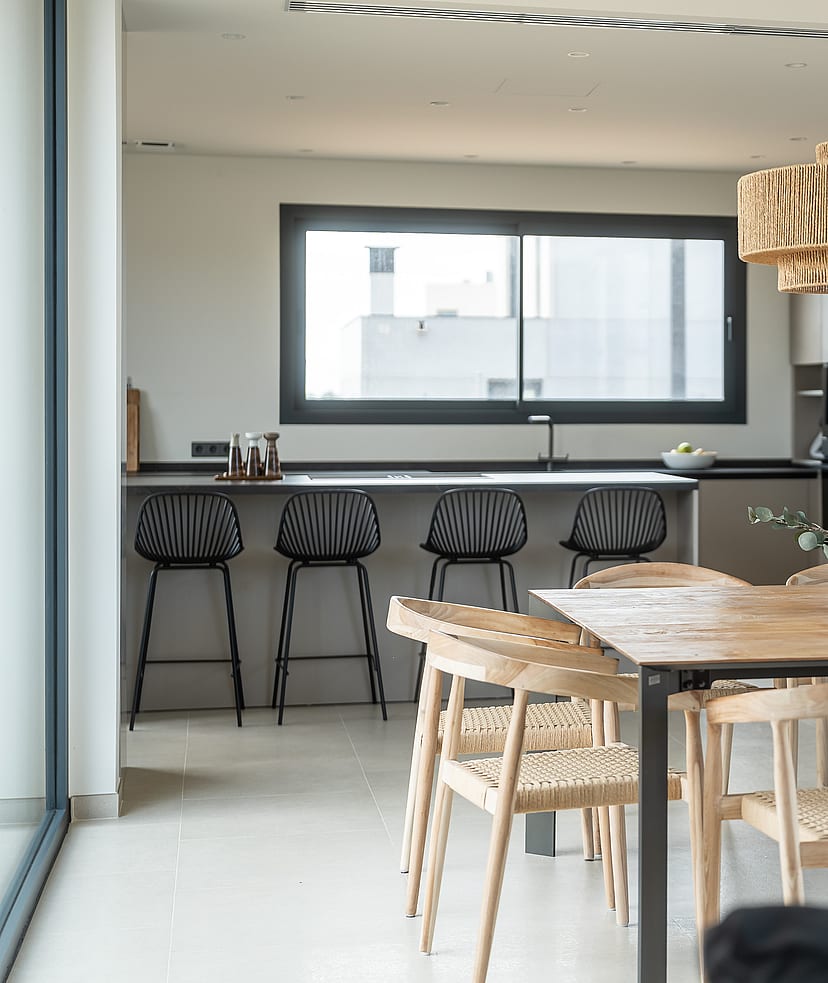 A dining space featuring a large square wooden table with woven chairs, a textured light fixture, and a modern kitchen island with black bar stools in the background.