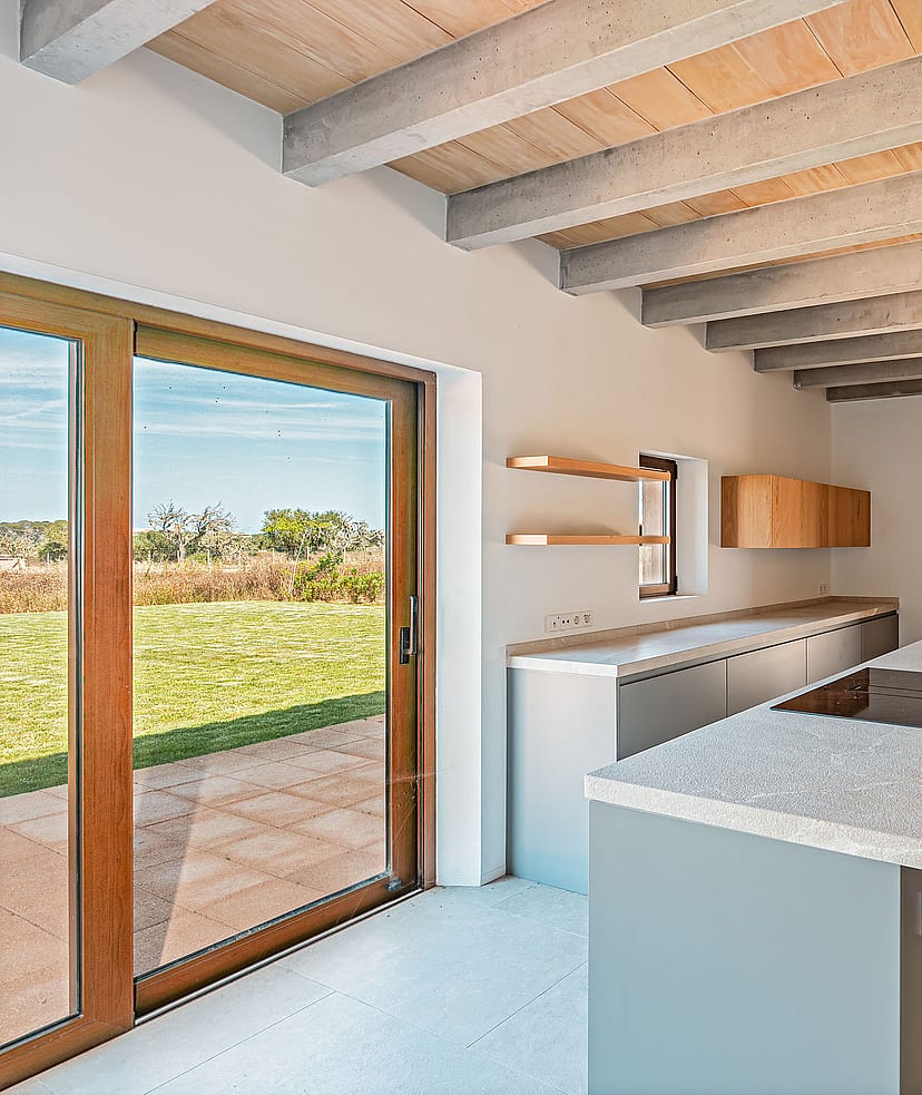 Modern kitchen featuring a central island, light grey cabinetry, wooden ceiling beams, and large glass doors overlooking a field.