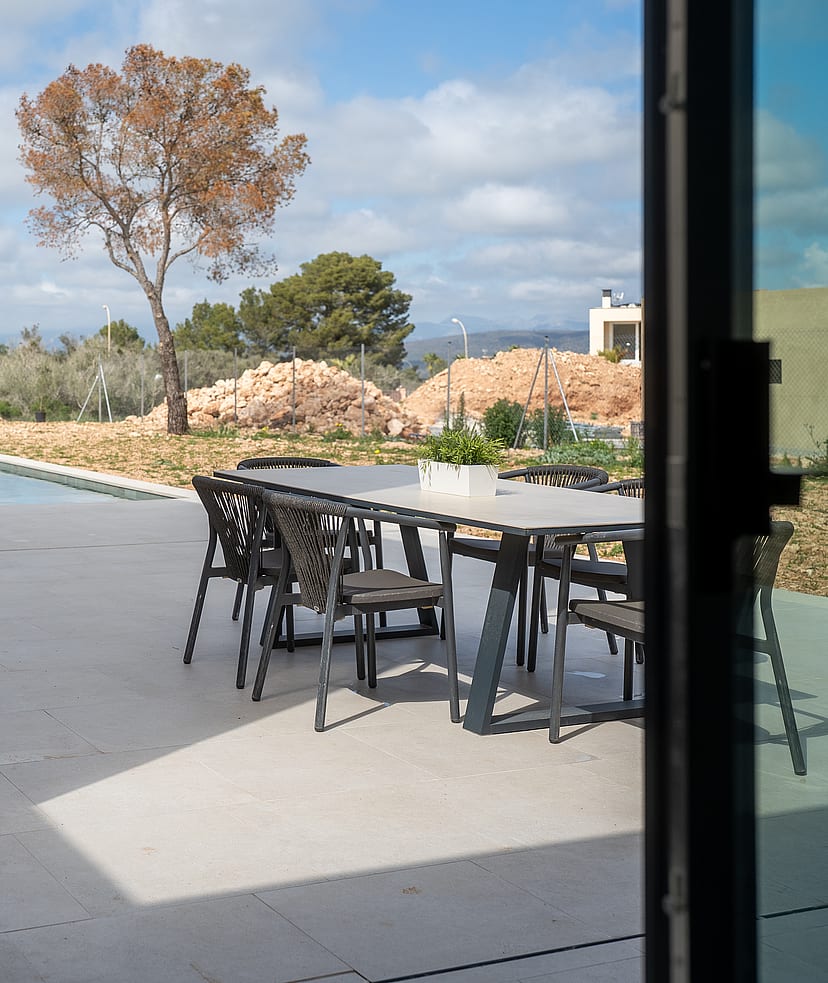 A covered terrace with a dining set and sun loungers overlooks a swimming pool and distant hills under a sunny sky.