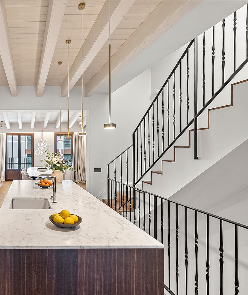 Kitchen island with marble top and dark wood base beside a staircase with black iron railings and exposed ceiling beams.