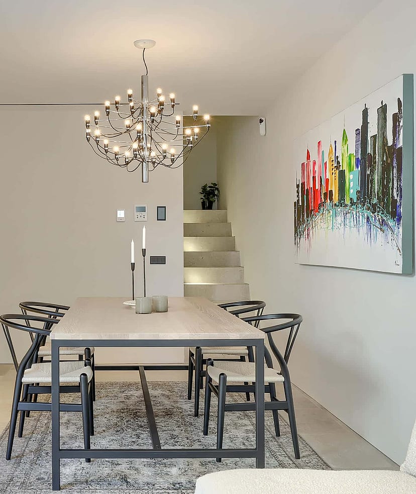 Interior view of a minimalist dining room with a stone table, black wooden chairs, a modern branching chandelier, and an abstract multi-colored painting on a white wall.