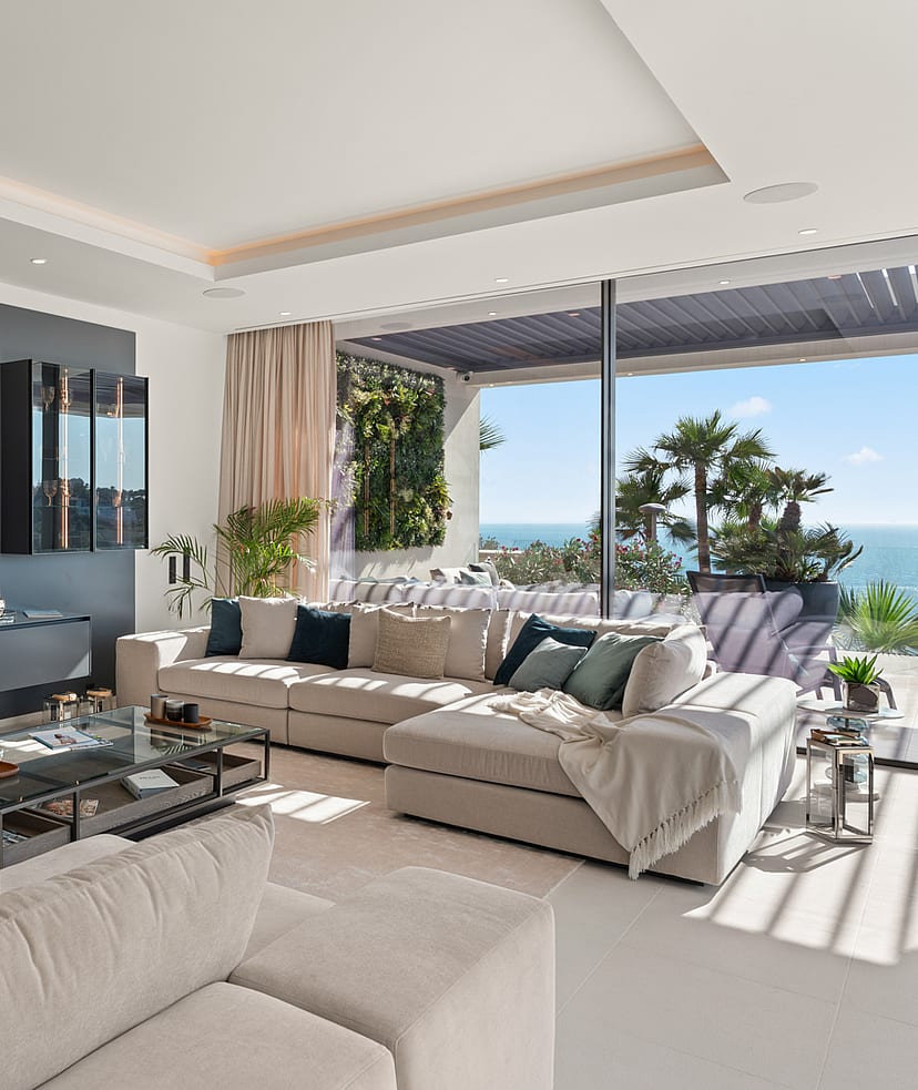Modern living room with a charcoal grey feature wall, a wall-mounted flat-screen television, a beige sectional sofa, and floor-to-ceiling glass sliding doors opening to a terrace with sea views.