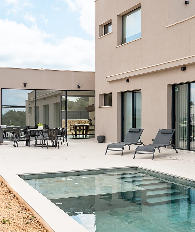 Rear facade of a modern beige villa showing a stone terrace, swimming pool with steps, and outdoor dining area.