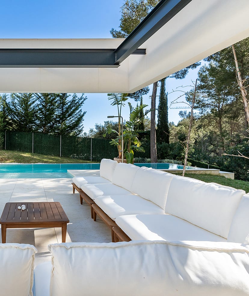 White cushioned outdoor seating under a steel pergola roof beside the swimming pool and pine garden