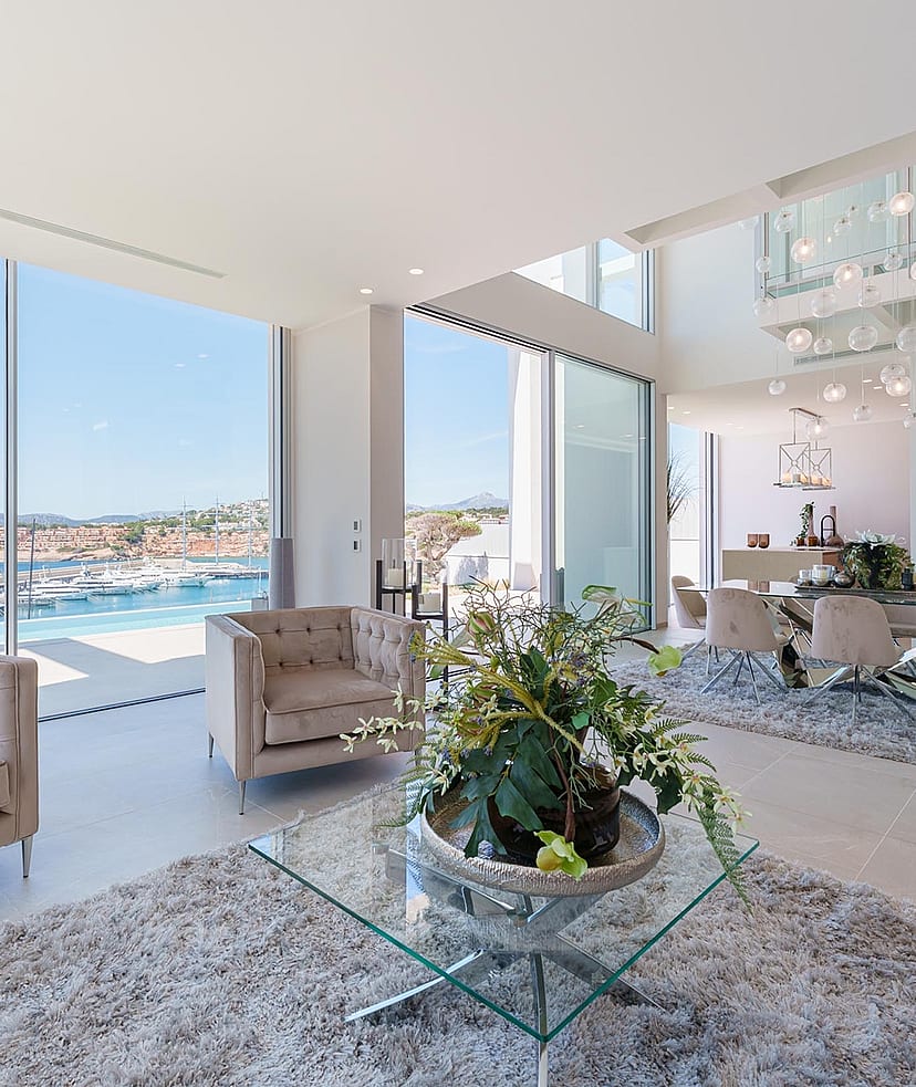 Modern open-plan living room with floor-to-ceiling windows overlooking Port Adriano marina, featuring tufted furniture.